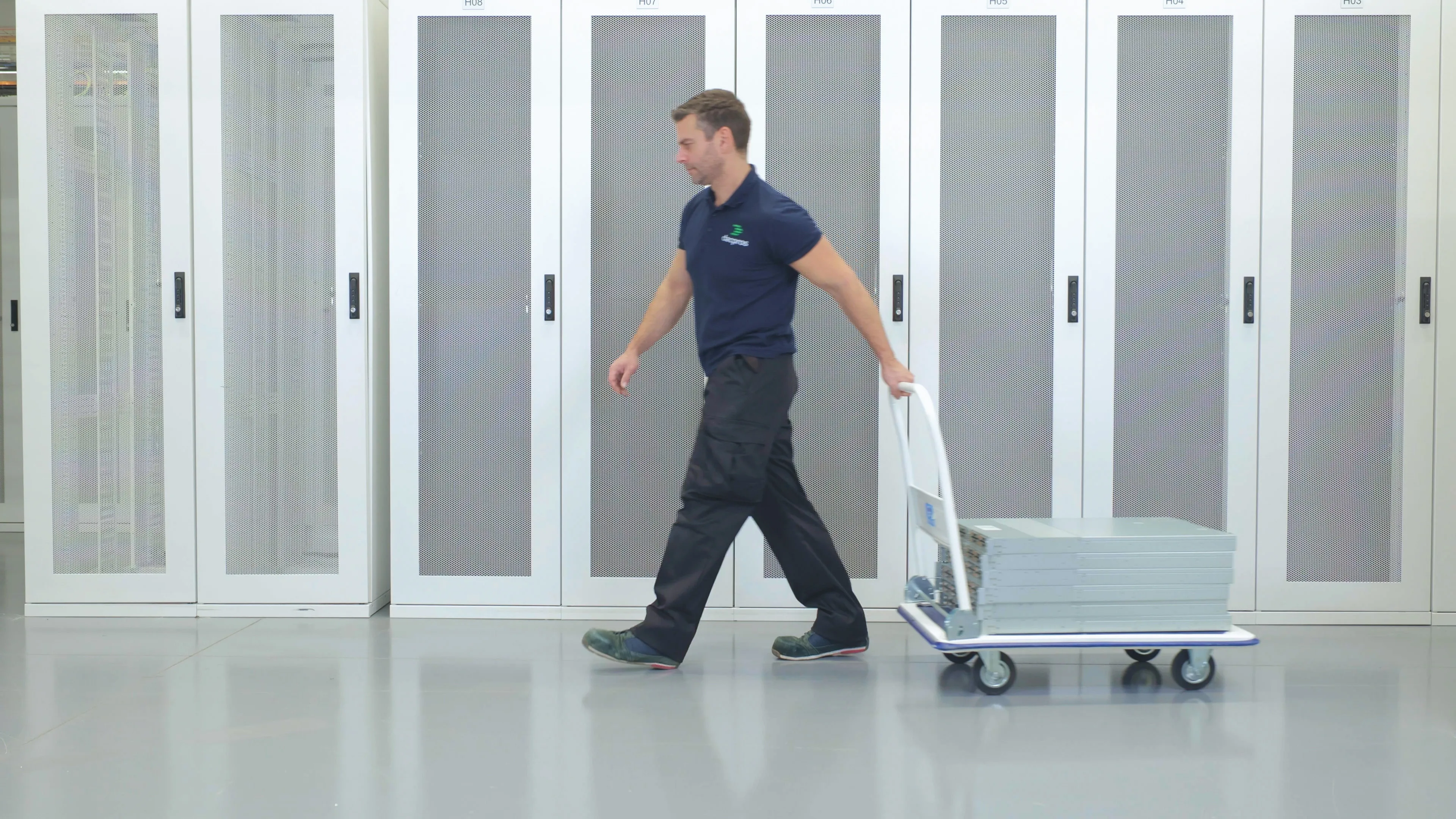 DACPROS engineer walking the data centre floor pulling a flatbed trolley past a row of server cabinets.