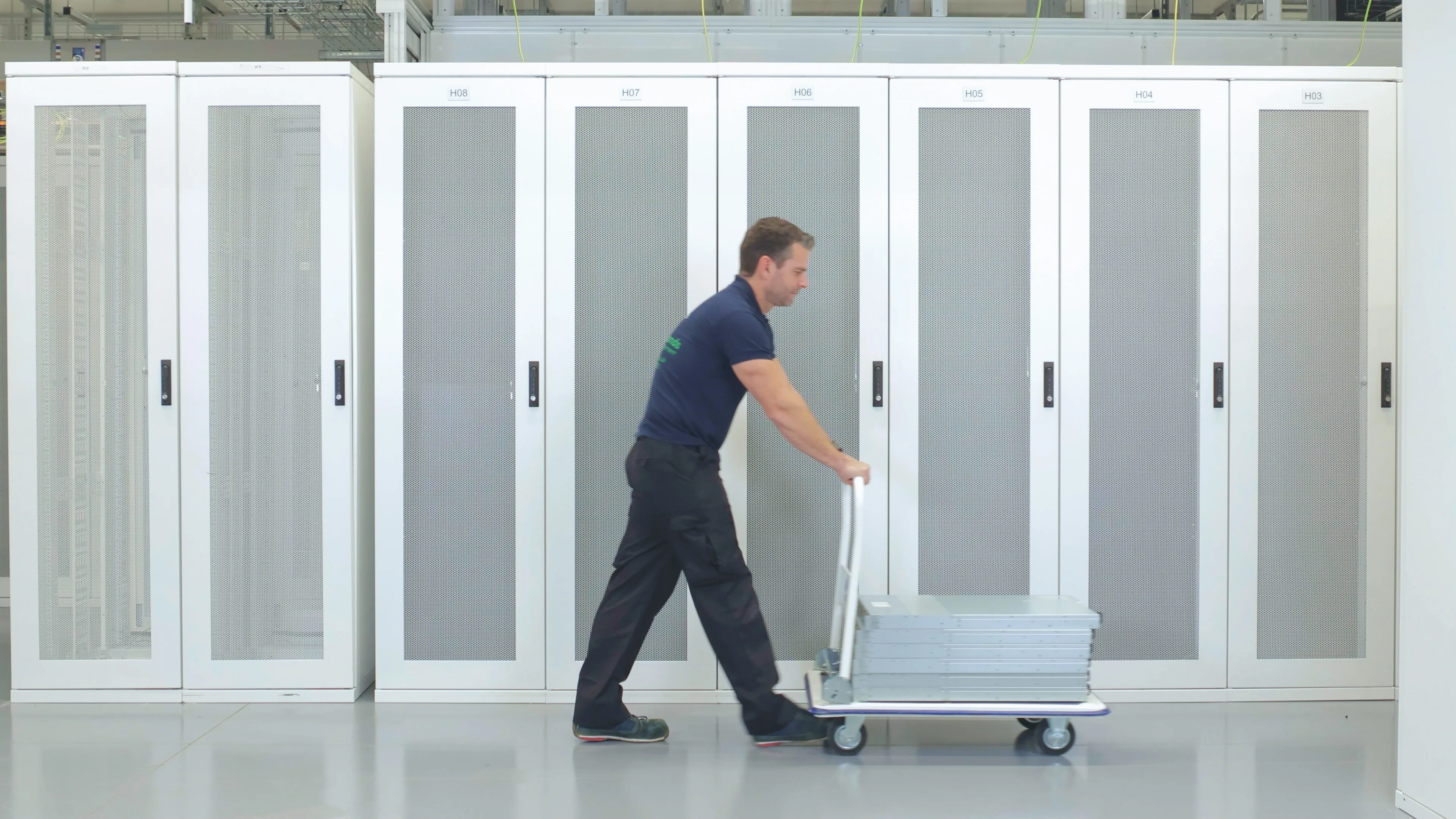 DACPROS engineer pushing equipment on a trolley along a row of server cabinets — the full hardware lifecycle in motion.